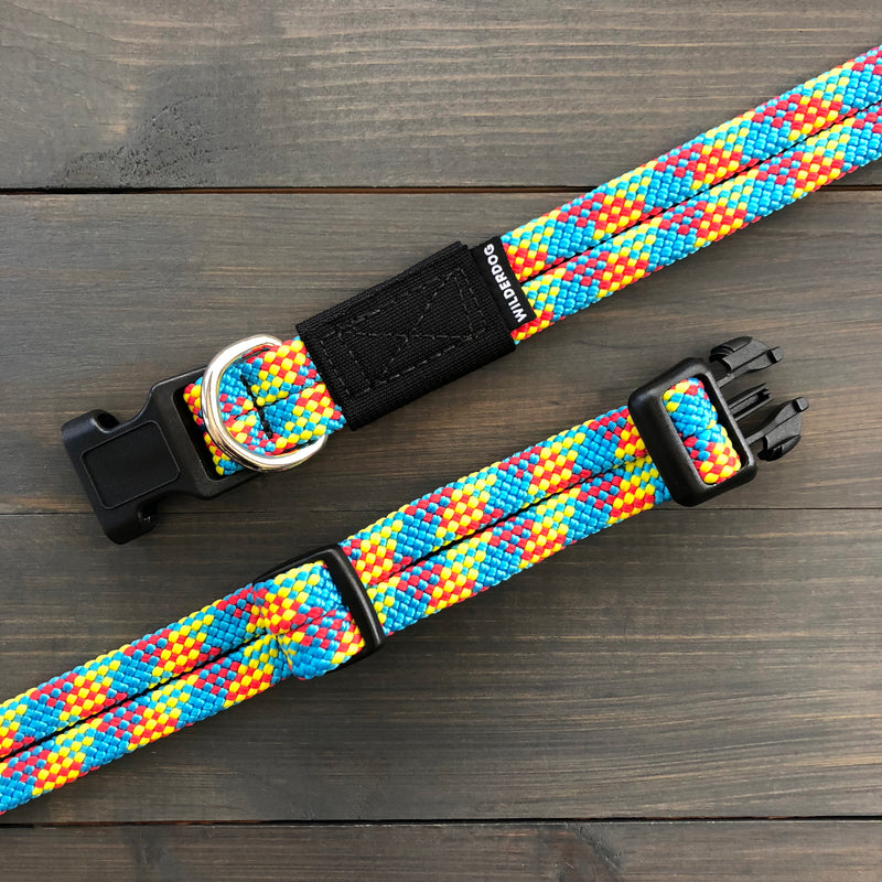 Two colorful, heavy-duty Aprés Collars with D-rings and adjustable plastic buckles lie on a dark wooden surface. One collar is fully extended while the other is fastened. The collars feature a vibrant pattern with blue, yellow, red, and green threads.